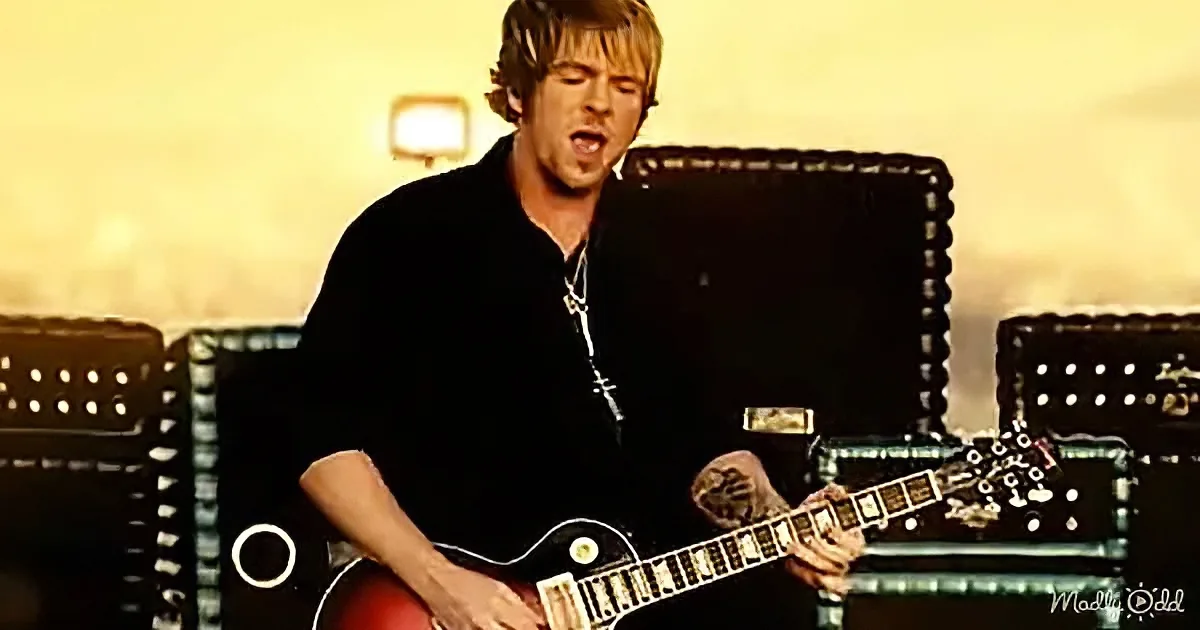 A man in a black shirt plays a red electric guitar on stage with amplifiers in the background.