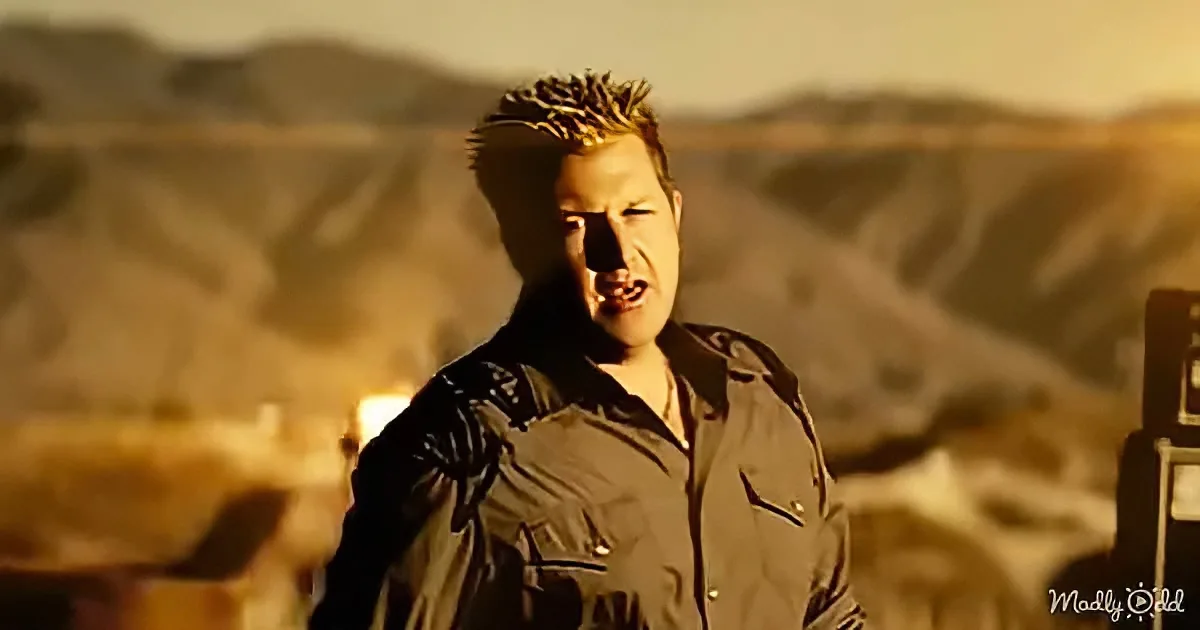 A man with spiky hair and a brown shirt stands in front of a desert landscape, mouth open.