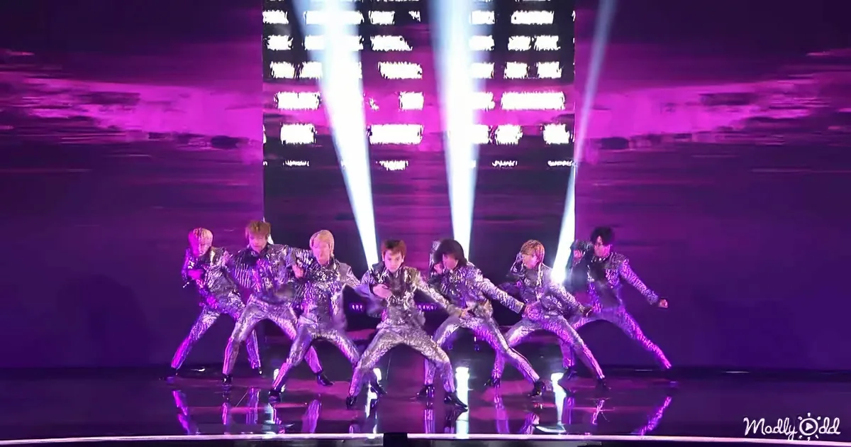 A group of dancers in shiny silver outfits performs a synchronized routine on a stage with purple lighting.