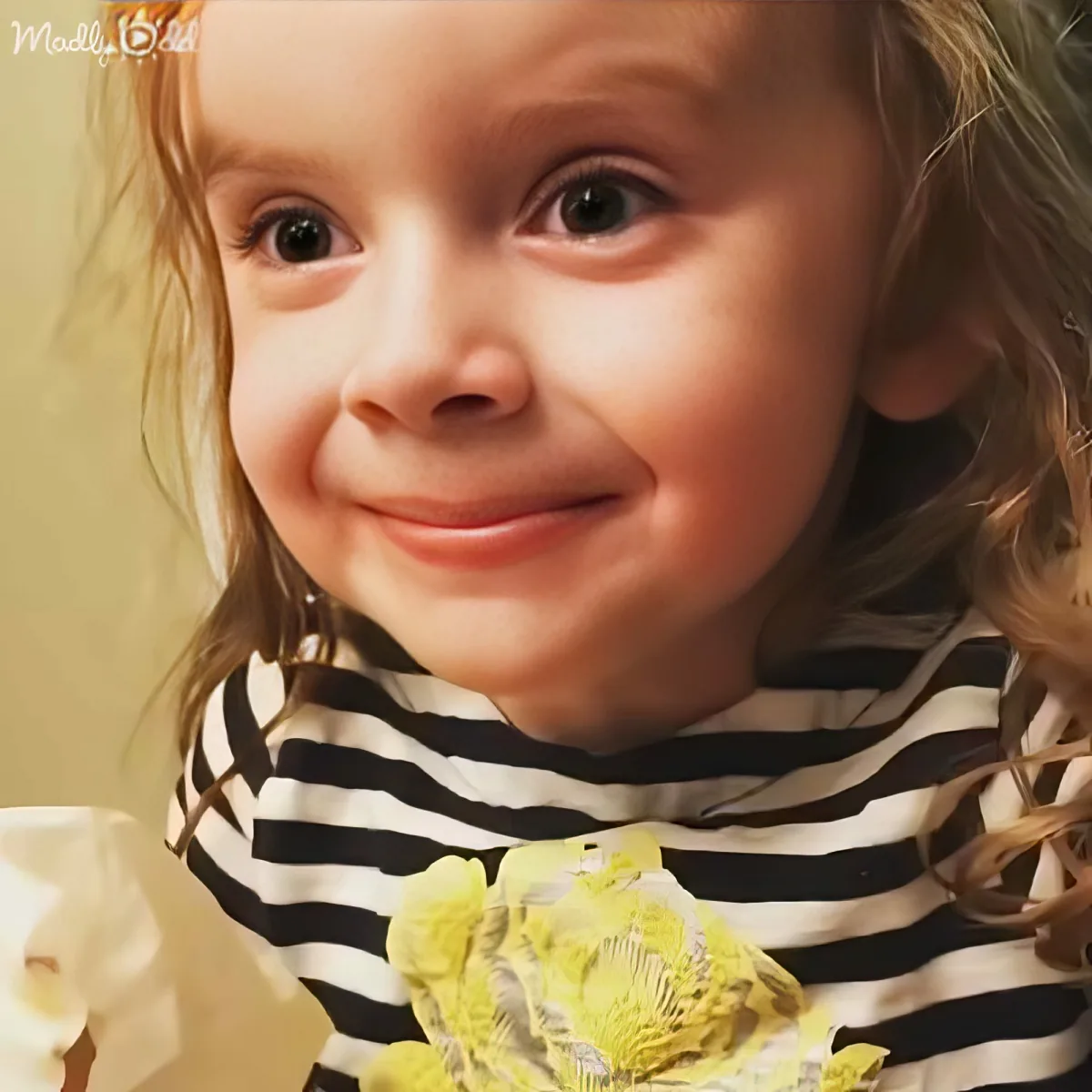 A young child with curly hair, wearing a black and white striped shirt with yellow accents, smiles softly.