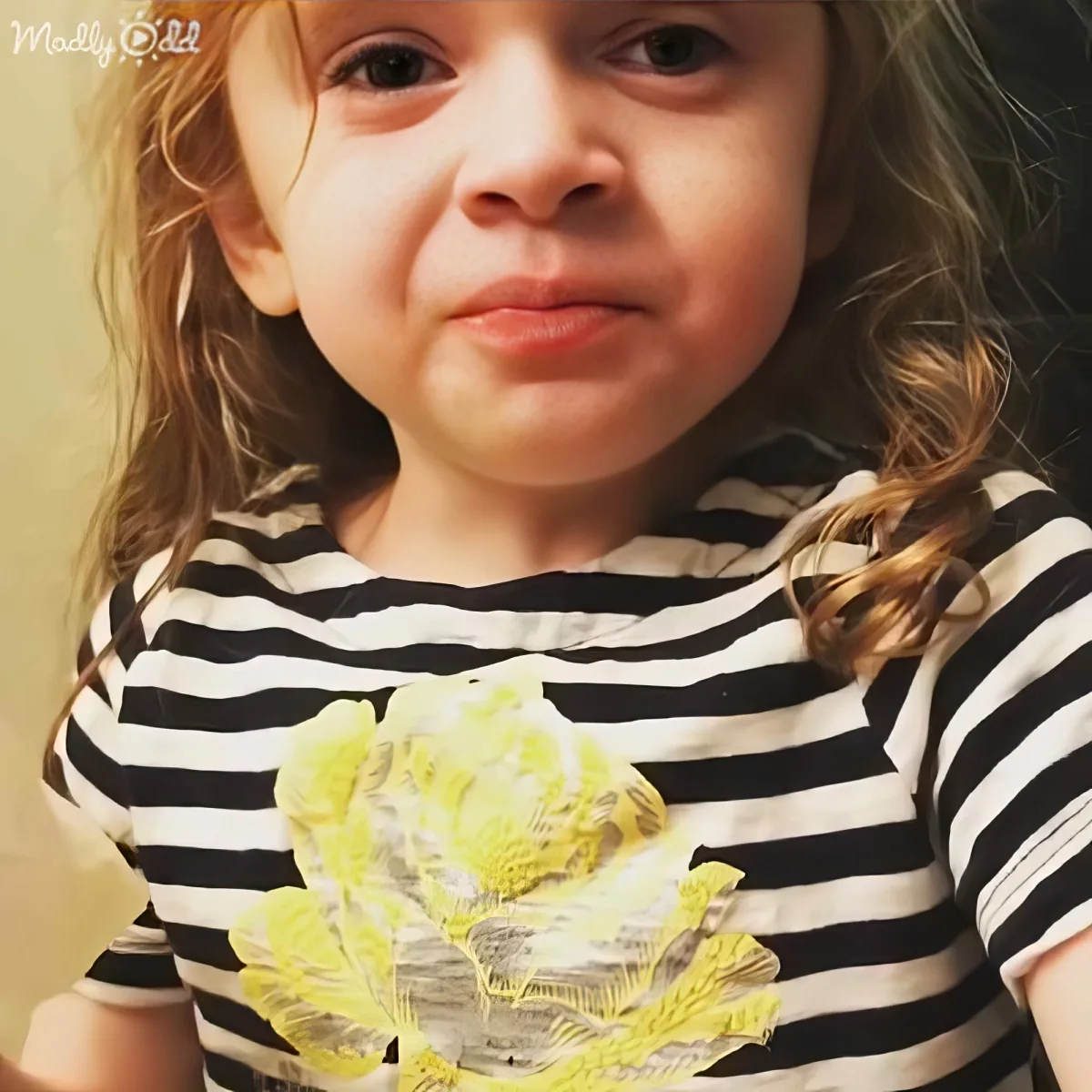 A young child with messy hair, wearing a striped shirt with a large yellow flower, looks directly at the camera.