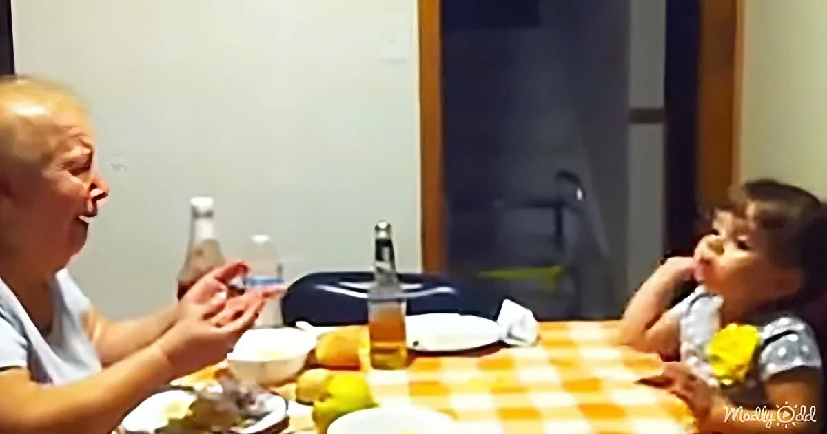 Two children sit at a checkered table, one clapping, the other looking up, with food and drinks around them.
