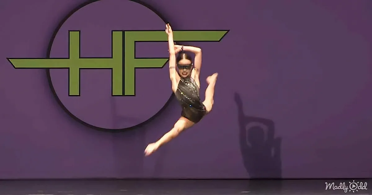A gymnast in a black leotard performs a split leap on stage with a "HF" logo in the background.
