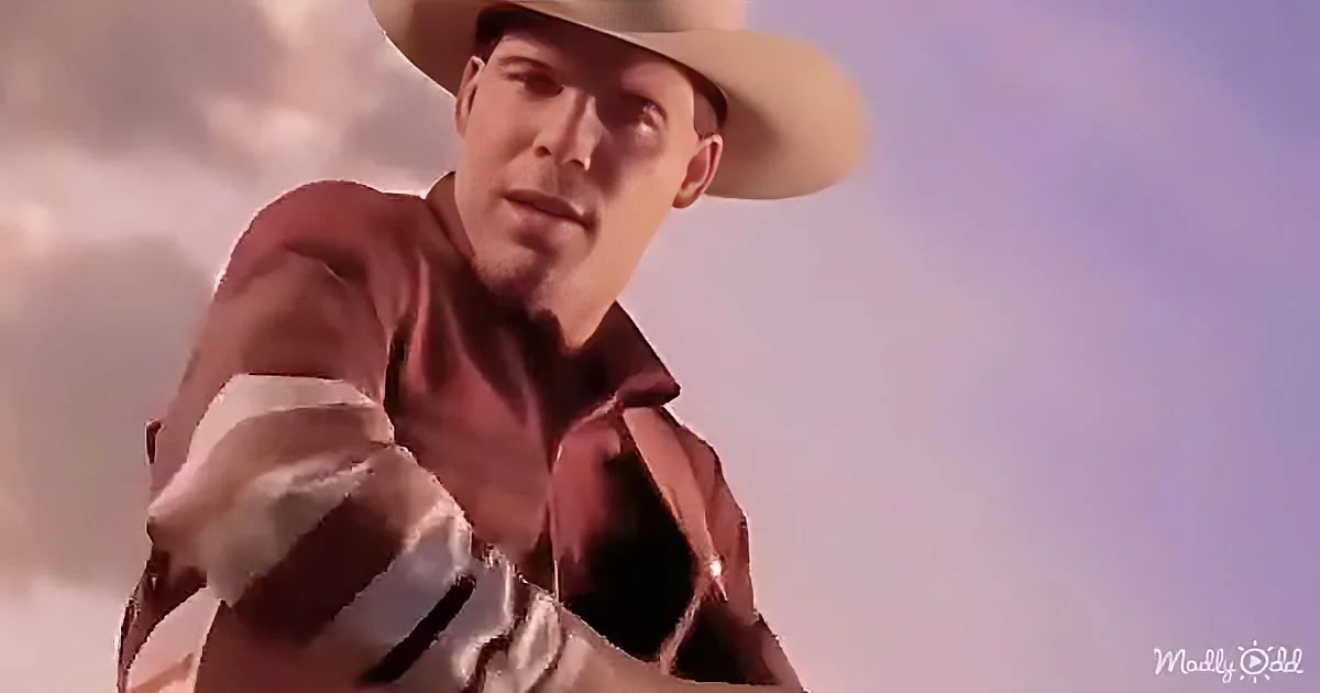 A cowboy in a red shirt and white pants, wearing a wide-brimmed hat, stands against a cloudy sky.