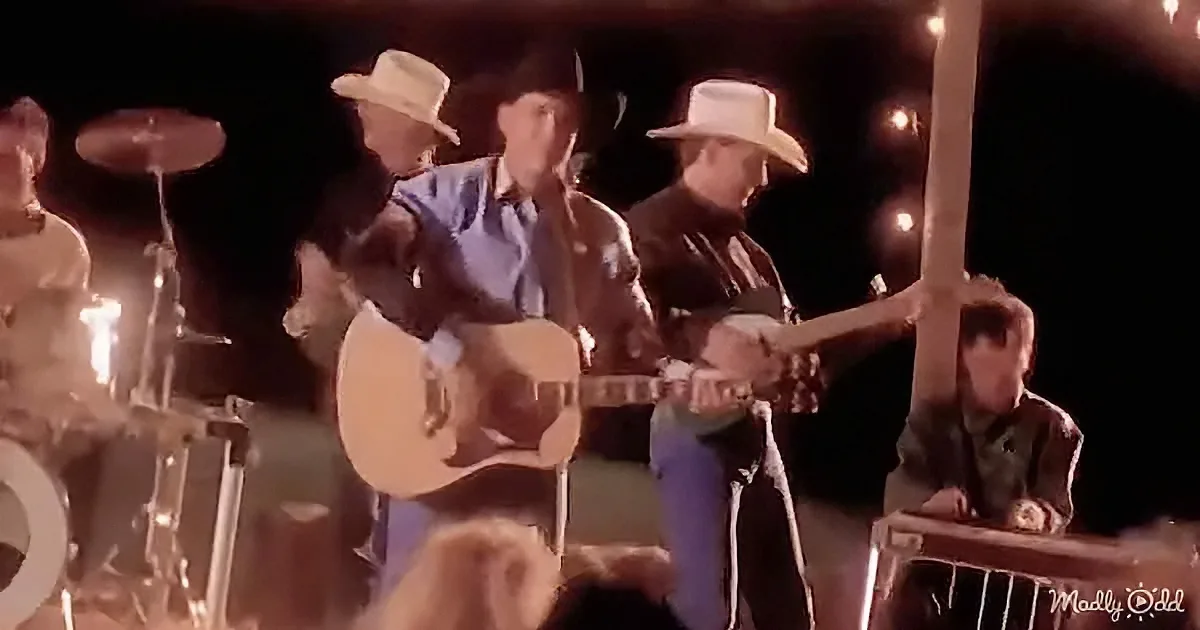A man in a cowboy hat plays guitar on stage with a band, surrounded by musical instruments and a dark background.
