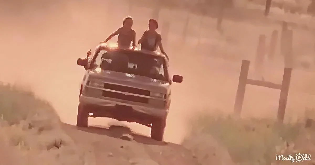 Two people ride on the back of a truck driving on a dusty road.