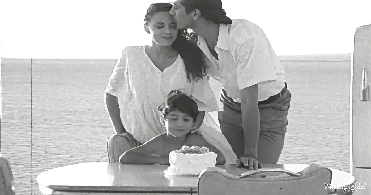 A woman and child sit at a table with a cake, while a man leans in to kiss the woman on the cheek.
