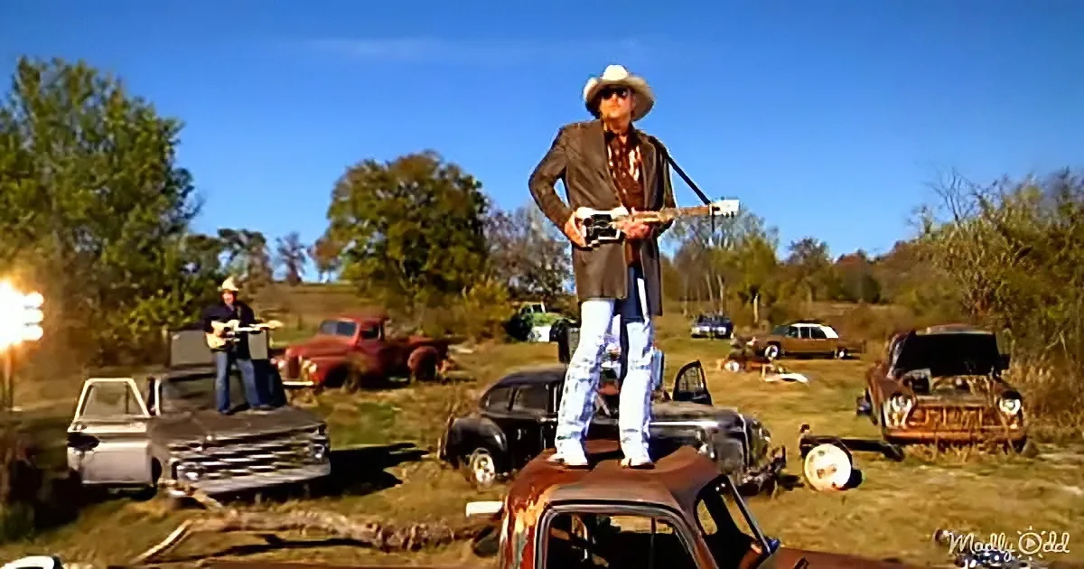 A man in a cowboy hat stands on a rusted car, holding a guitar, surrounded by vintage vehicles in a field.