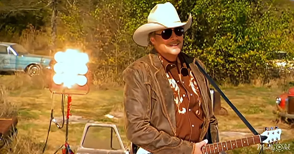 A man in a cowboy hat and sunglasses plays guitar outdoors, with a vintage car and a bright light in the background.
