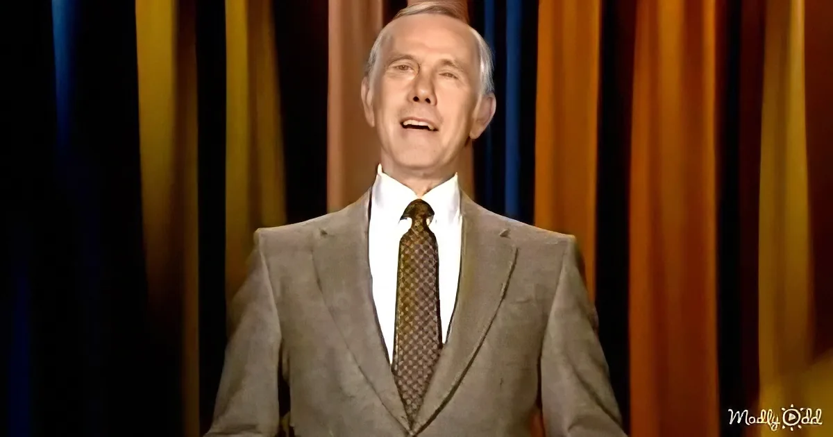 A man in a suit and tie stands in front of a colorful curtain, mouth slightly open.