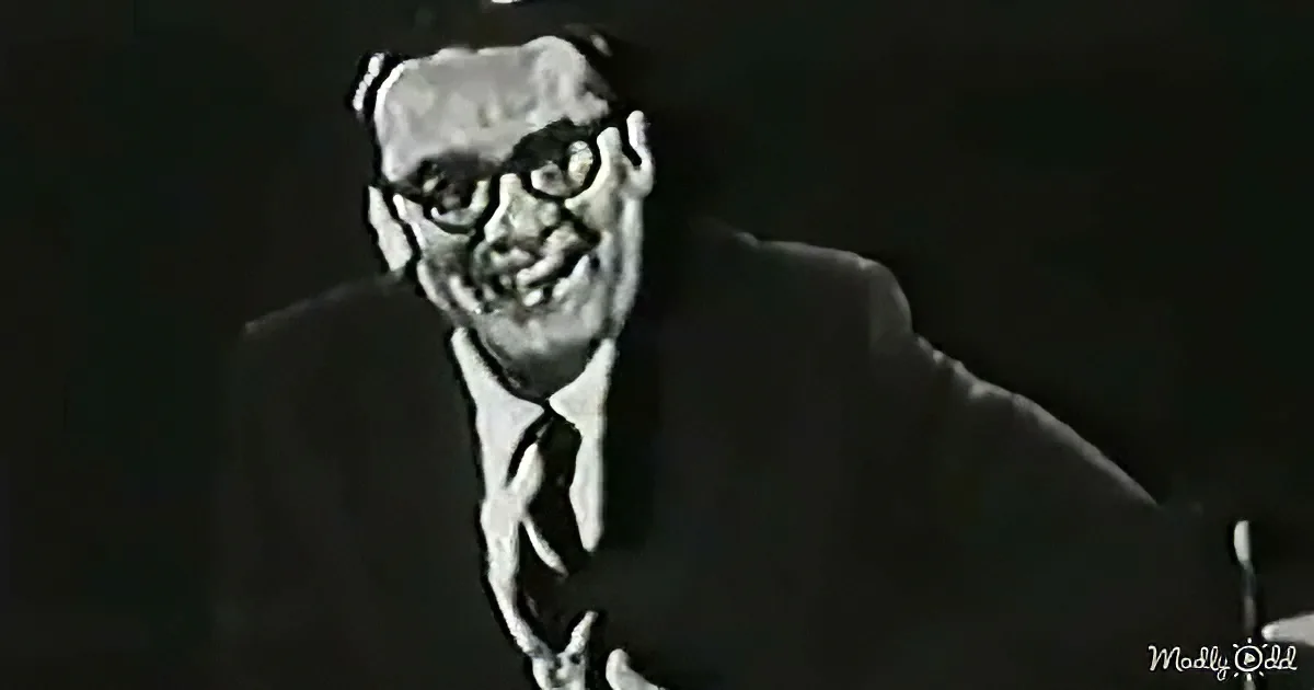 A man in a suit and tie, wearing glasses, smiles while holding a microphone.