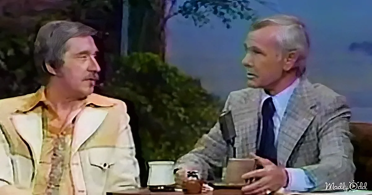 Two men are seated at a table, one gesturing while the other listens, with a backdrop of trees and a microphone.