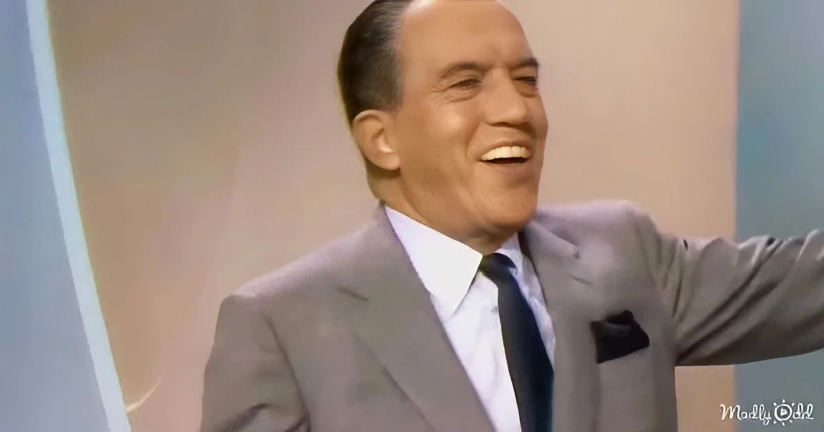 A man in a gray suit and tie is smiling and raising his arm, possibly in a celebratory gesture.