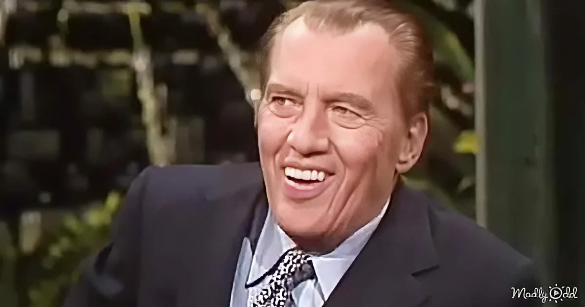 A man in a suit and tie smiles widely, with a blurred background of greenery and structures.