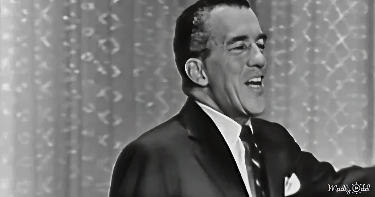 A man in a suit and tie is speaking into a microphone, smiling, against a patterned backdrop.