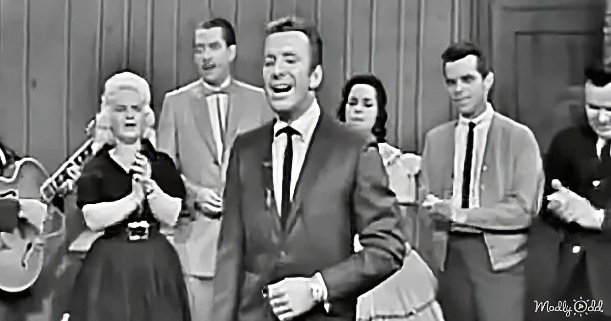 A man in a suit and tie is singing passionately, surrounded by a group of people clapping and smiling.