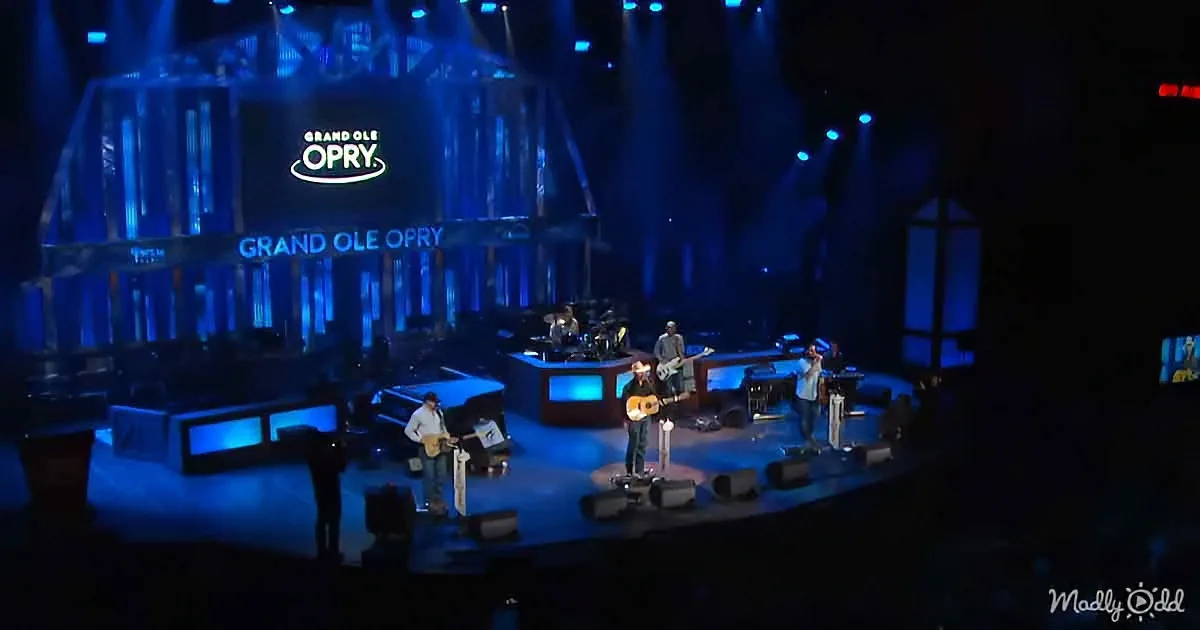Musicians perform on a stage at the Grand Ole Opry with blue lighting and a large sign in the background.