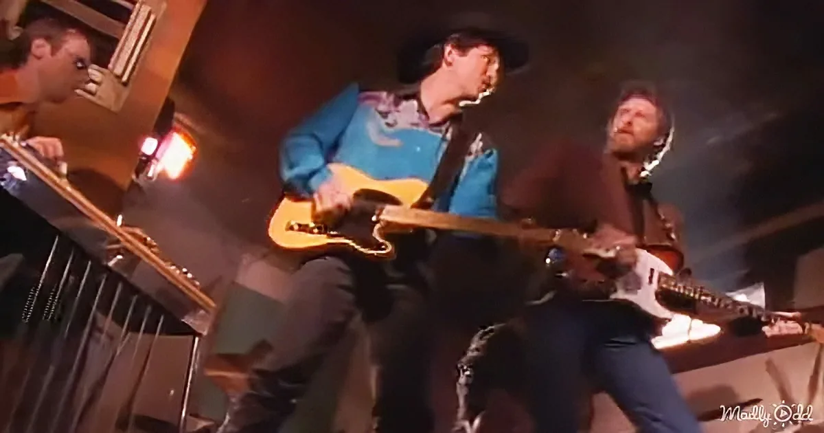 A man in a cowboy hat plays a guitar on stage, surrounded by other musicians.