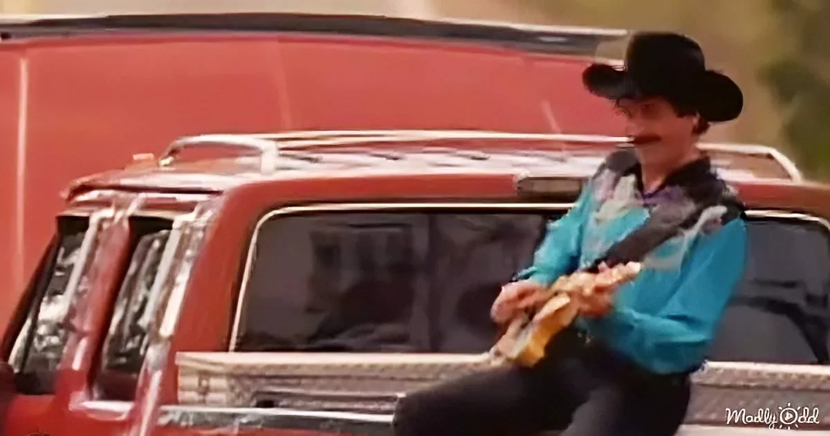 A person in a cowboy hat and blue shirt plays a guitar in front of a red truck.