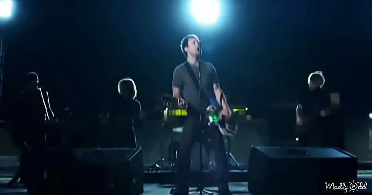 A man in a dark shirt stands on stage, illuminated by a bright light, surrounded by other musicians.