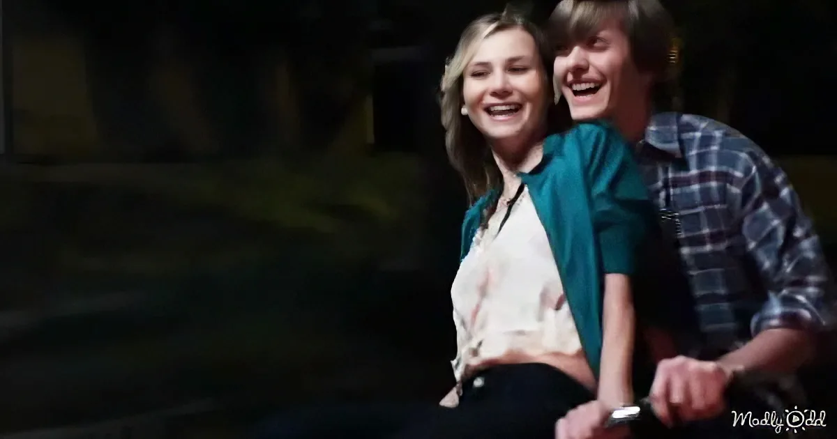 Two people, one in a green top and white shirt, the other in a plaid shirt, are laughing together in a dimly lit room.
