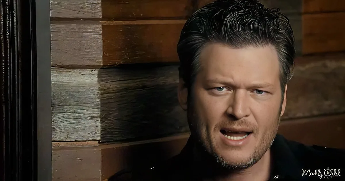 A man with short dark hair and a beard, wearing a black shirt, is speaking in front of a rustic wooden background.