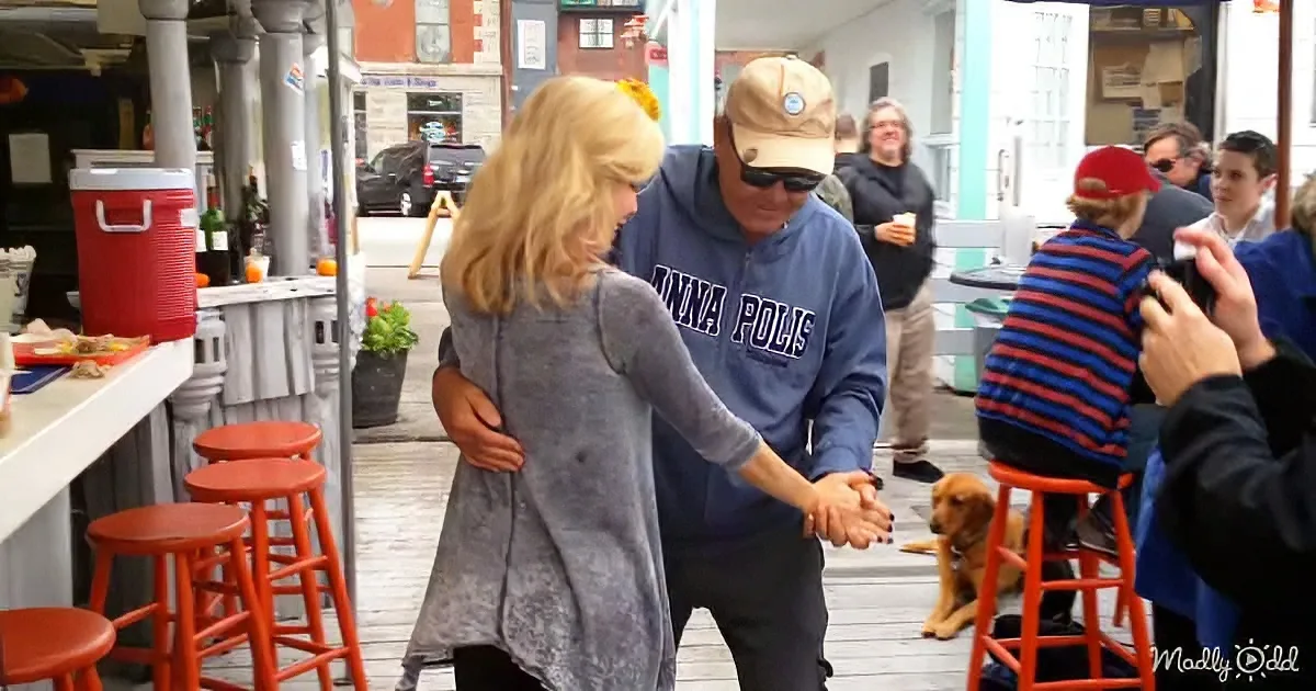 A man in a "Minneapolis" hoodie and woman in a gray coat are dancing on a wooden deck with a dog nearby.