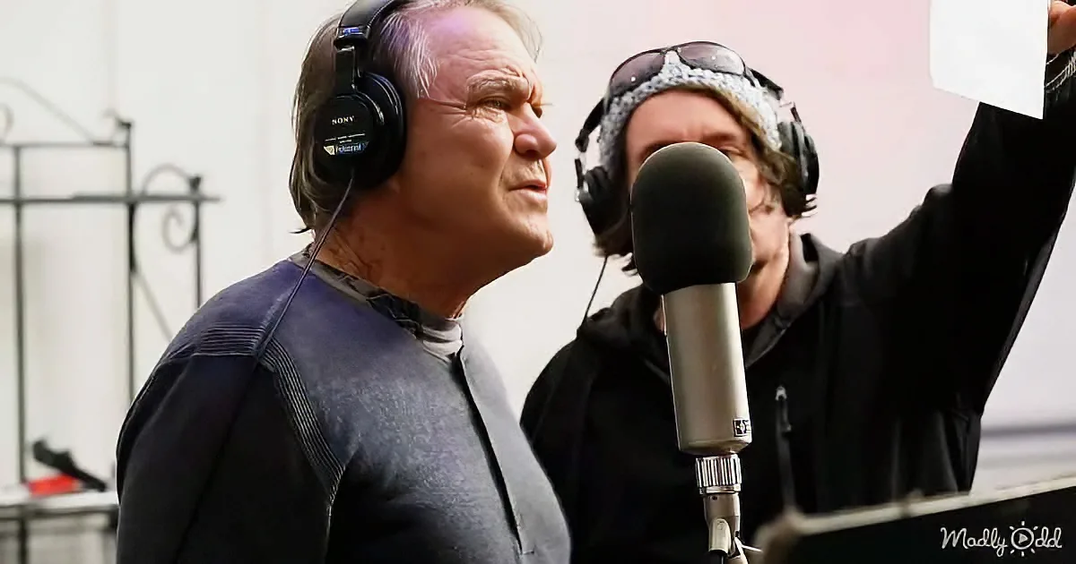An older man wearing headphones stands in front of a microphone, with another person holding up a sheet of paper behind him.