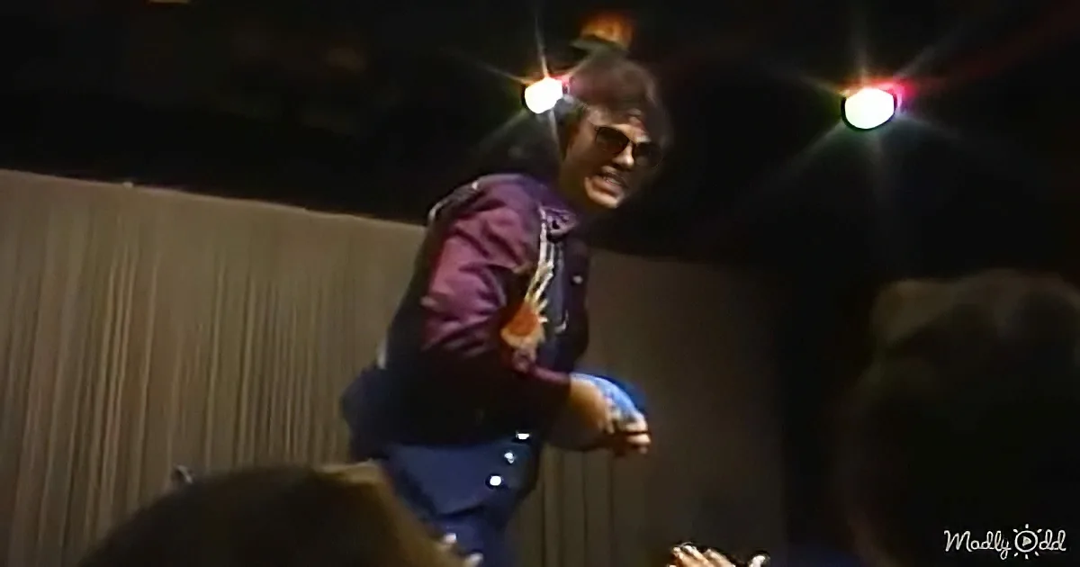 A man in a purple jacket and sunglasses is performing on stage, holding a guitar, with colorful lights above him.