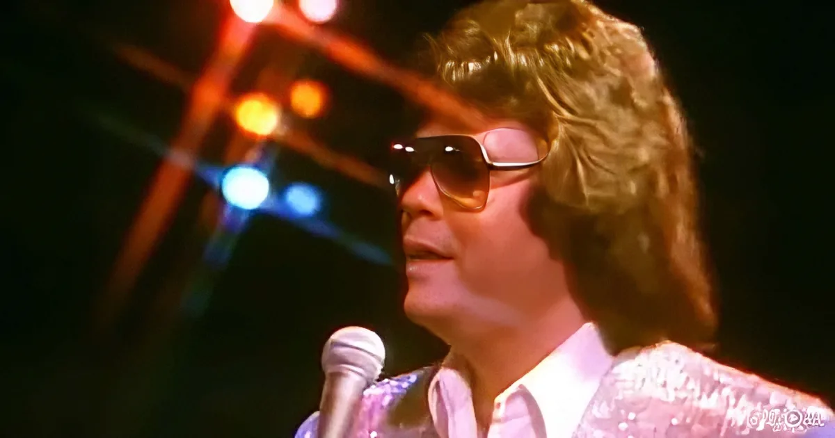 A person with curly hair and sunglasses sings into a microphone on stage with colorful lights.