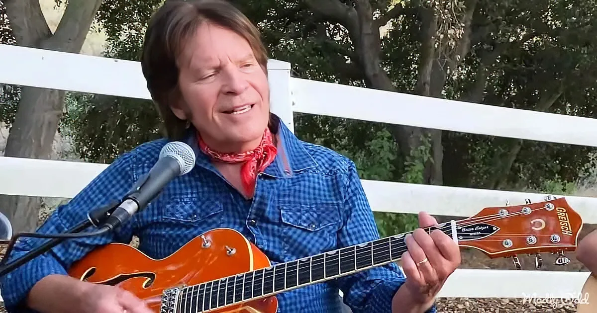 A man in a blue plaid shirt plays a Gretsch guitar, singing into a microphone outdoors.