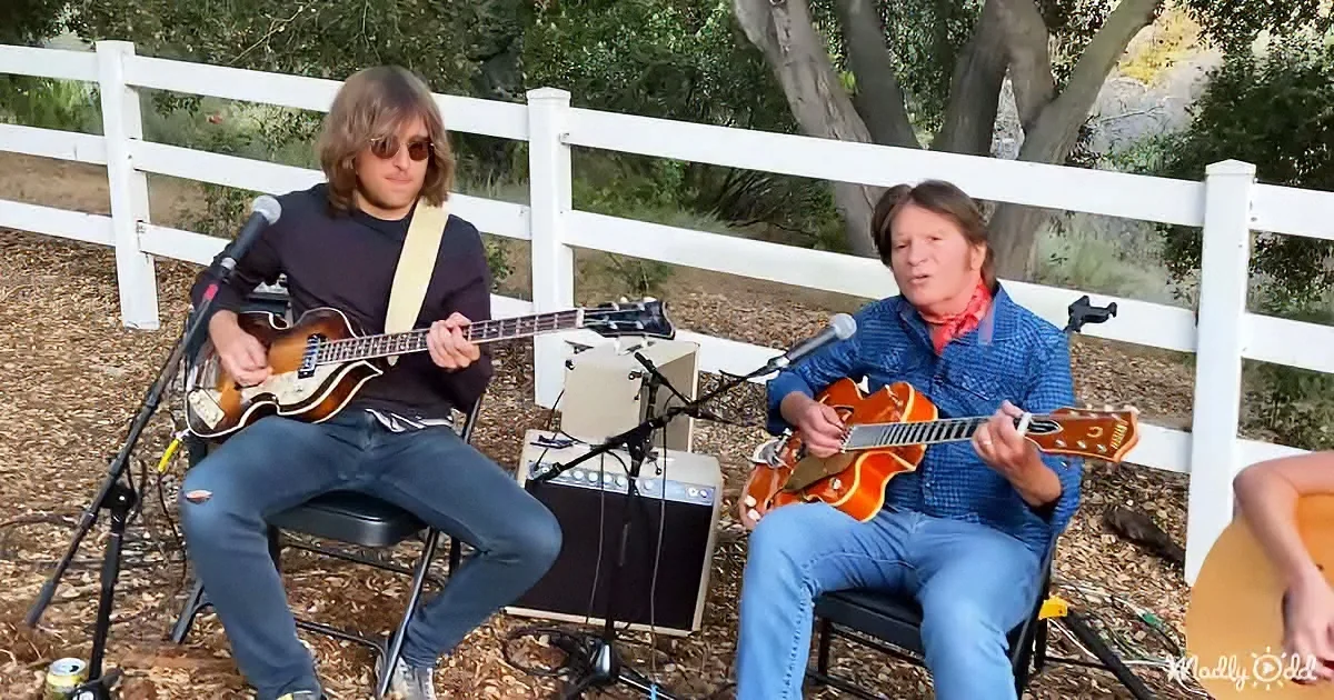 Two musicians play guitars outdoors, one with a bass, surrounded by microphones and amplifiers.