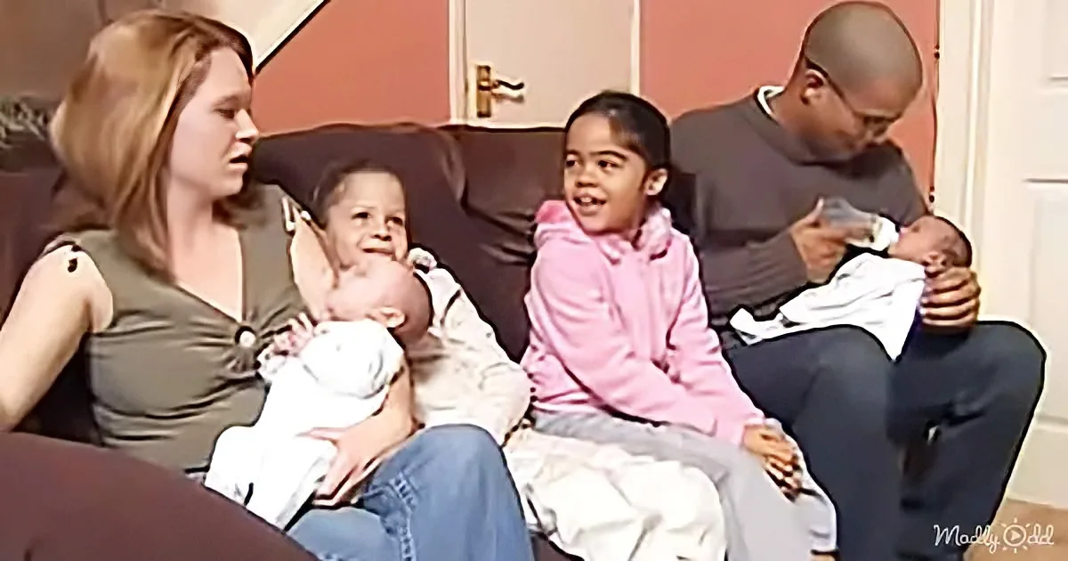 A family of five, including two adults and three children, is sitting together on a couch, with two babies being held and...