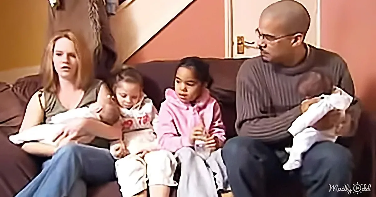 A family of five sits on a couch, each holding a baby, in a cozy living room setting.