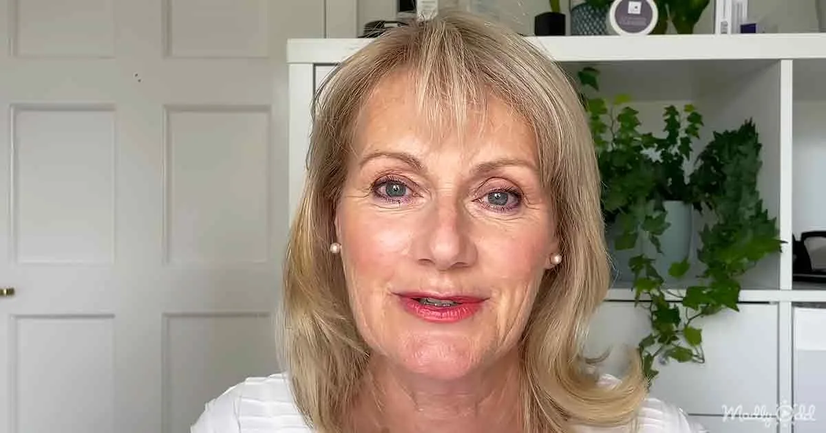 A woman with blonde hair and pearl earrings smiles in a room with a white door and green plants.