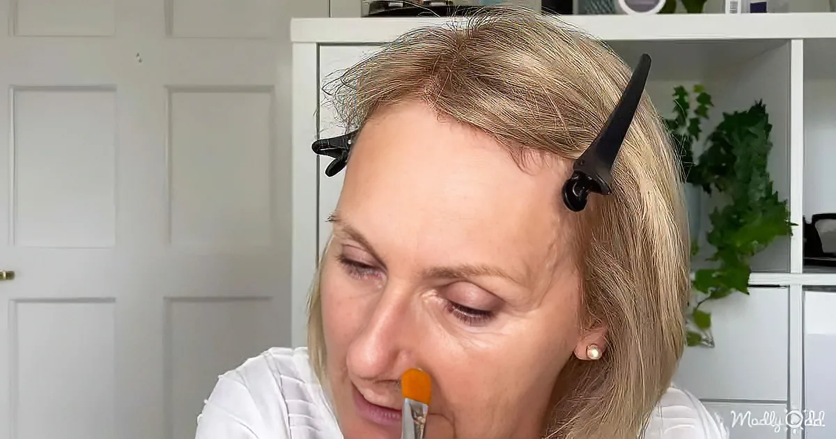 A woman with blonde hair and a white shirt is applying makeup, with hair clips holding her hair back.