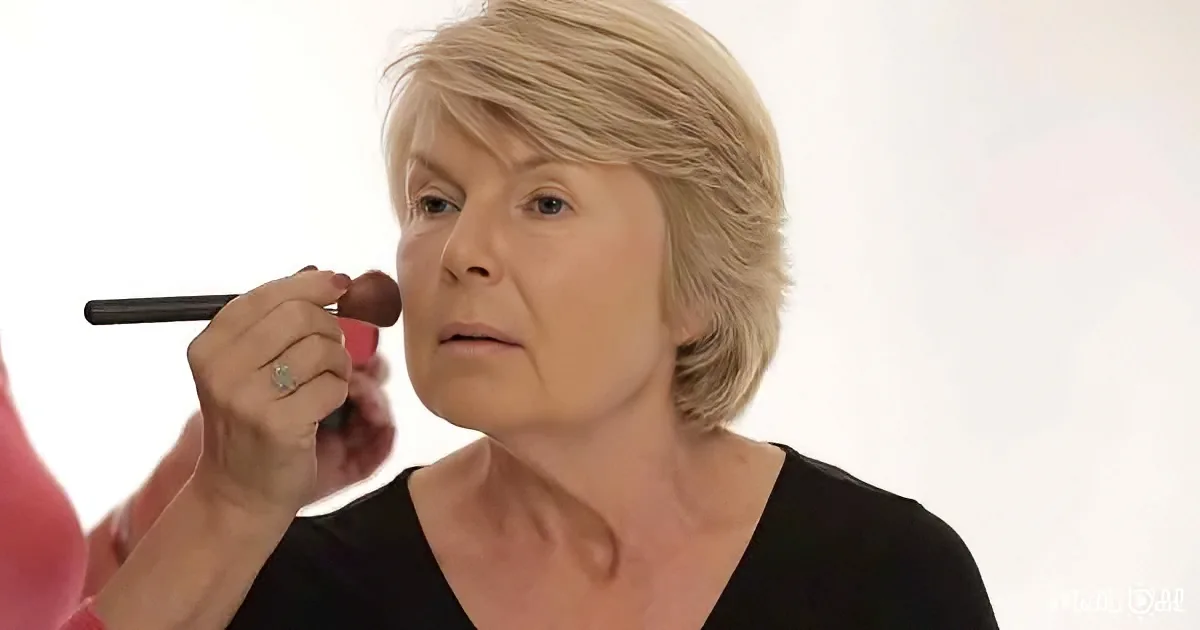 An older woman with short blonde hair is having makeup applied to her face.