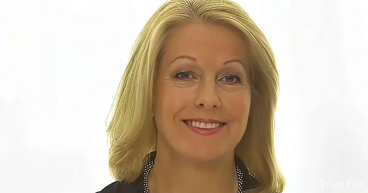 A smiling woman with blonde hair, wearing a black top and pearl necklace, against a plain white background.