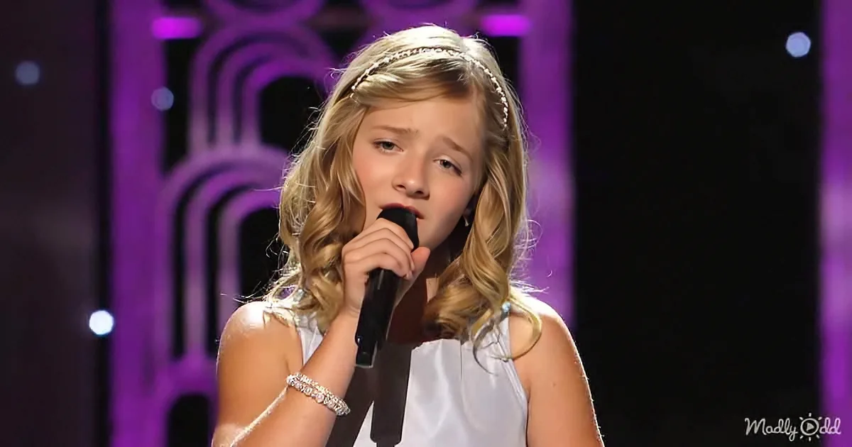 A young girl with blonde hair, wearing a white dress and a headband, sings into a microphone on stage with a purple...
