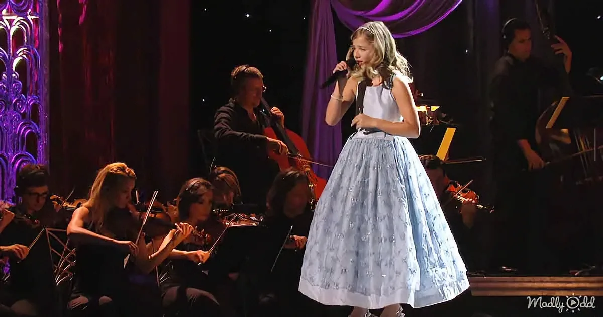 A young girl in a light blue dress sings on stage with an orchestra in the background.