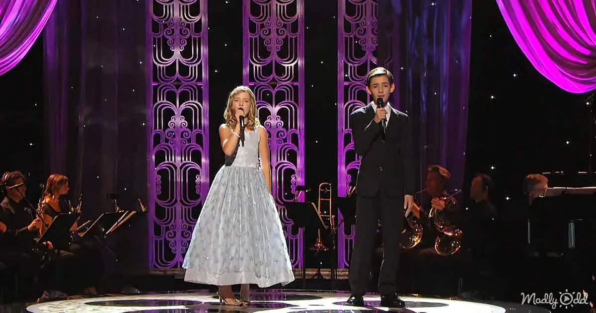 A young girl in a silver dress and a boy in a black suit sing on stage with a purple backdrop and orchestra.