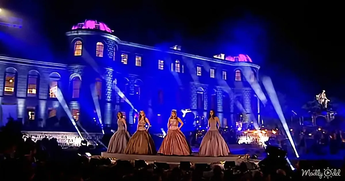 Four women in pink dresses stand on stage in front of a grand building illuminated by blue lights.