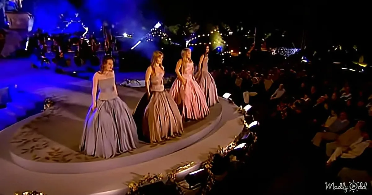Models in elegant dresses walk on a runway at a fashion show with an audience and stage lighting.