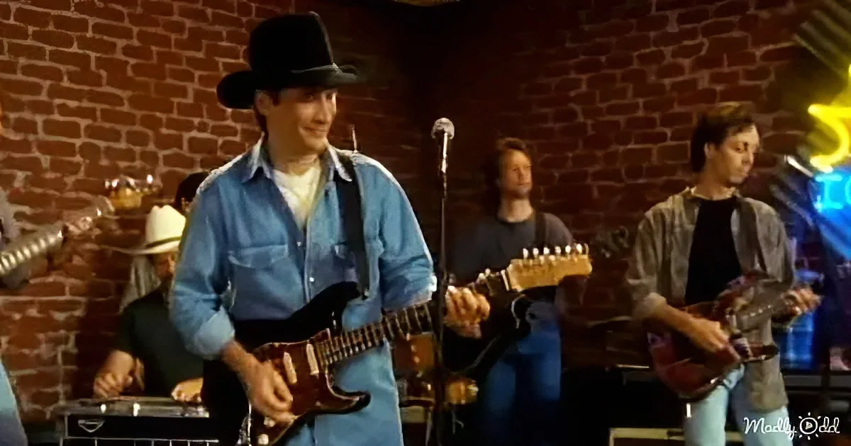 A man in a blue shirt and black hat plays guitar on stage with a band in a brick-walled venue.