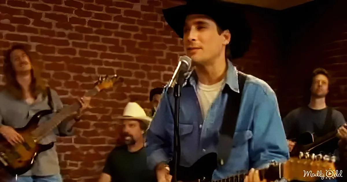 A man in a cowboy hat sings into a microphone, surrounded by musicians in a brick-walled room.