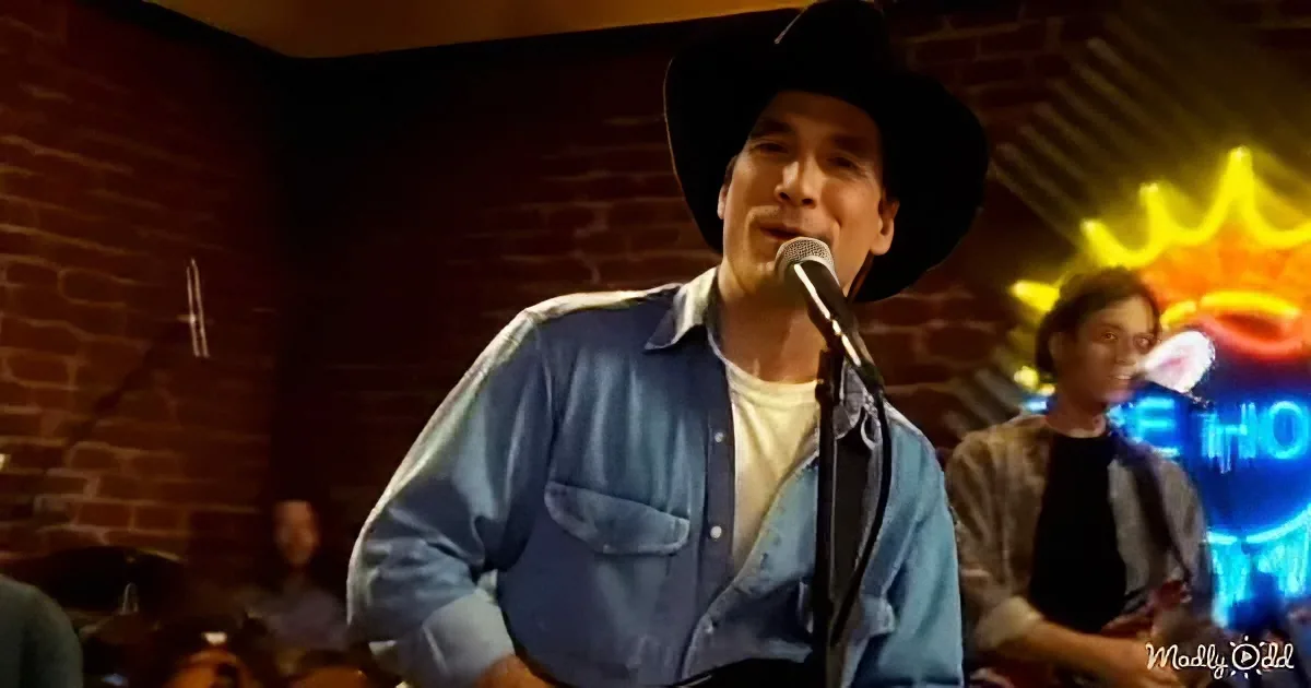 A man in a cowboy hat sings into a microphone, wearing a blue shirt, in a dimly lit bar with a neon sign.