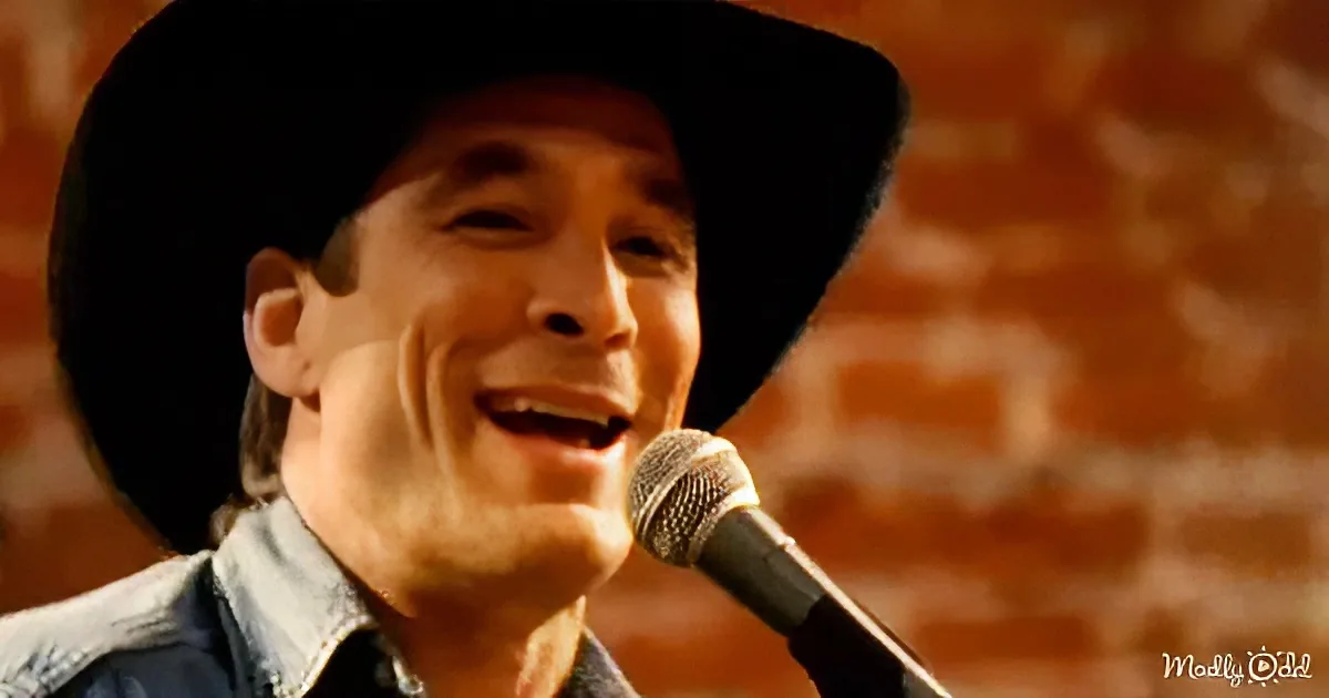 A man wearing a cowboy hat smiles while singing into a microphone against a brick wall.