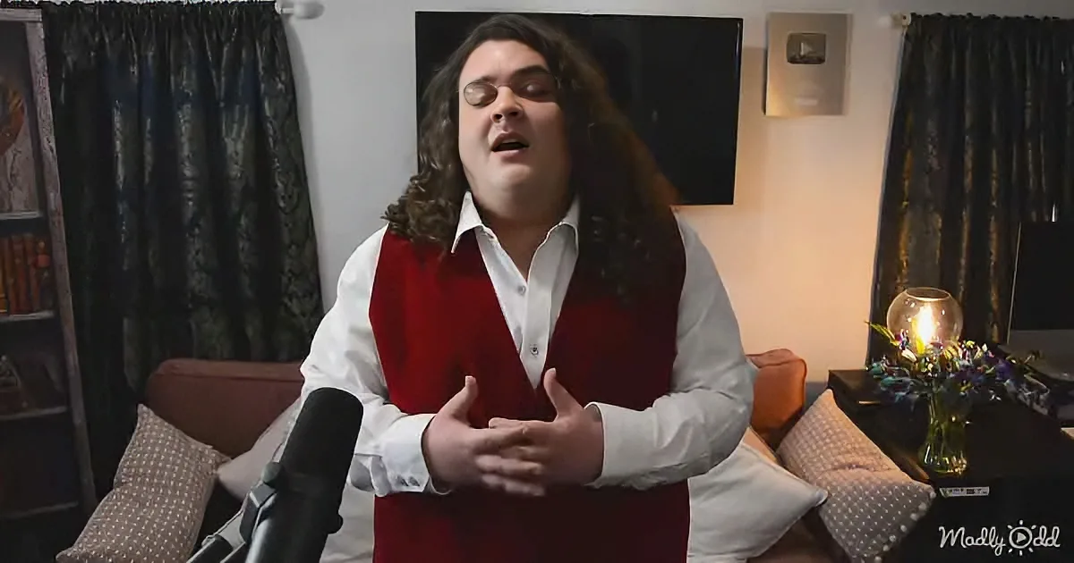 A man with long hair and a red vest sings into a microphone in a cozy room with curtains and a TV.