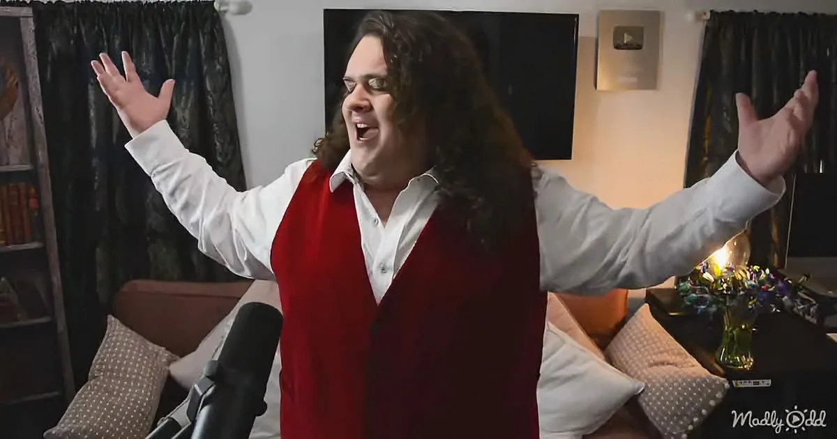A man with long hair, wearing a white shirt and red vest, stands with arms raised in a living room.