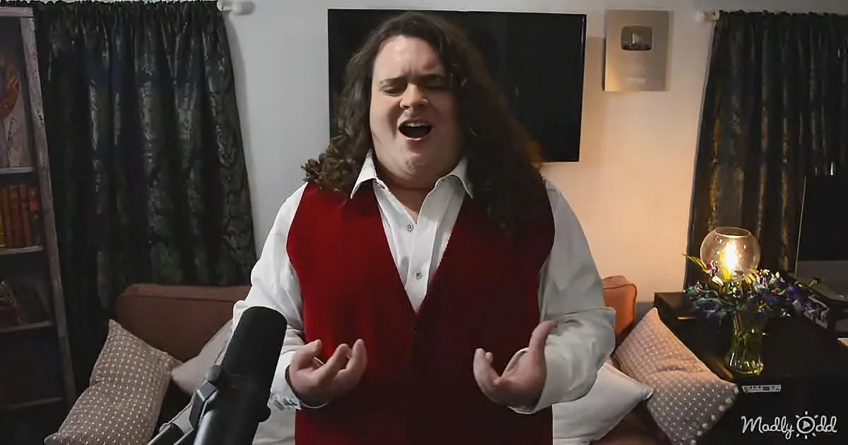 A person with long hair, wearing a red vest and white shirt, sings into a microphone in a cozy room with curtains and a lamp.